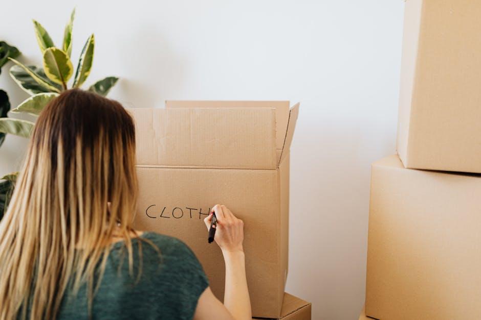 A woman with long, light brown hair is seen from behind, writing the word 'CLOTHES' on the side of a large, open cardboard box using a black marker. The scene takes place indoors during a home relocat
