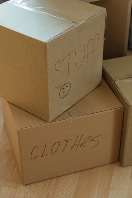 A woman with long, light brown hair is seen from behind, writing the word 'CLOTHES' on the side of a large, open cardboard box using a black marker. The scene takes place indoors during a home relocat