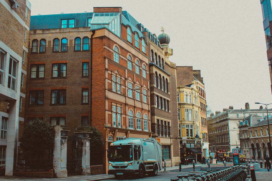 A small white moving truck with red markings parked on a wide urban street in Mayfair, London, with a large historic stone building with numerous arched windows visible in the background. To the left,