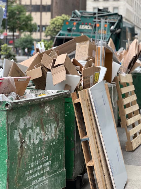 A side view of a small waste collection truck parked on a residential street during daytime, with the truck loaded with various disposal materials. The truck's cargo area is filled with cardboard boxe