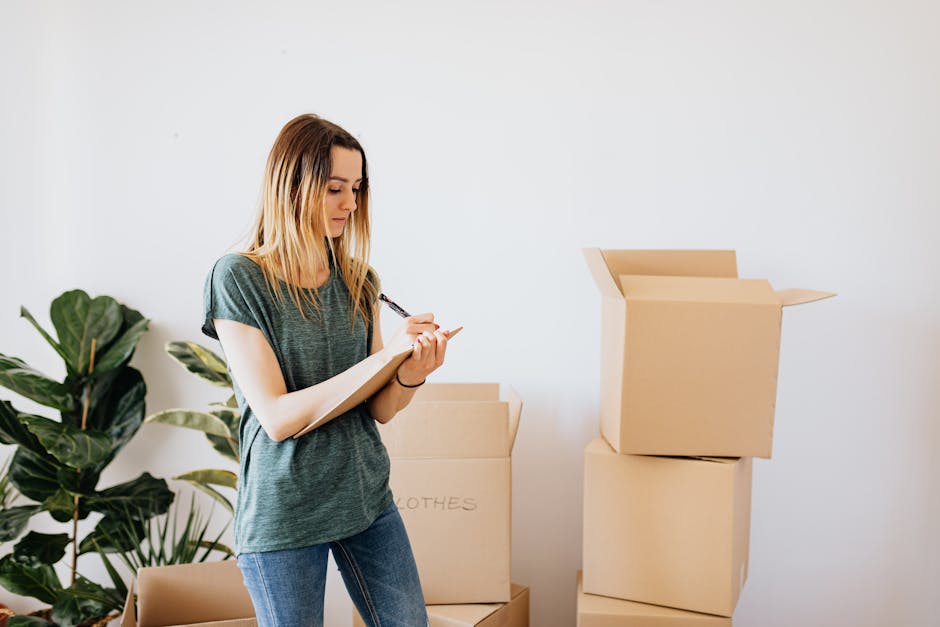A person in light-colored clothing is seen arranging cardboard boxes on a wooden floor inside a room, with sliding glass doors and natural light in the background. The individual is holding a mid-size