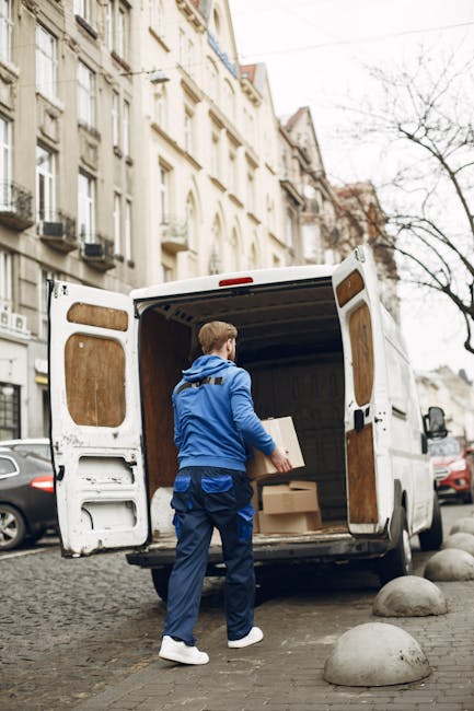 A male mover wearing a blue jacket, blue trousers, and white shoes is standing at the open rear doors of a white cargo van parked on a cobblestone street in an urban residential area. The van's interi